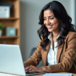 Smiling professional woman in a brown blazer working on a laptop in a modern office setting.